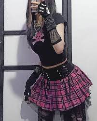 Pessoa com look gótico/punk: camiseta preta com estampa de caveira rosa, saia xadrez rosa e preta, luvas rendadas e acessórios escuros